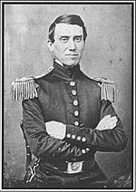 Black and white photo of a clean-shaven man with deep-set eyes and dark hair. With arms folded, he wears a dark uniform with two epaulettes.