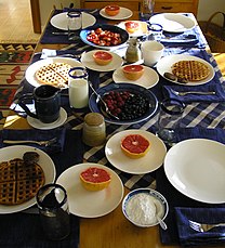 Table covered in mugs, glasses of milk, and plates of waffles, berries, powdered sugar, and grapefruit.