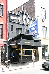 Street view in broad daylight of a two-story city building with a piano-shaped awning, gold-framed window panels at the bottom floor and jazz musician icons toward the roof, and a flag stemming from the top floor with the club's name; two pedestrians walk outside the building.