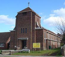 A red brick church stands with the small steeple facing the street as the church's entrance. The building extends back from the entrance, and the building is surrounded by grass.