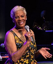 A grey-haired dark-skinned woman in a brightly patterned dress singing into a microphone