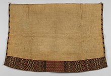 A photo of a woven cloak laid flag. It is mostly off-white, with geometric designs along its left, right, and bottom borders.