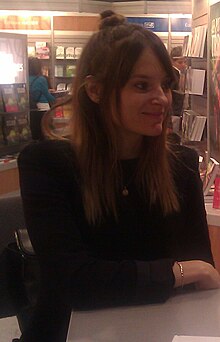 blurry image of Stéphanie Lapointe wearing a black top, seated at a table in what appears to be a bookstore, smiling and looking left of camera