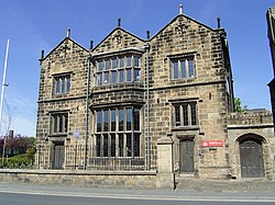 Dark stone building with three ranges and many tall mullioned windows close to and facing the road