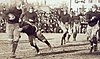 An old, grainy black and white photo showing a college football game from 1919. A player is running with the ball on the left side of the picture with several players in pursuit to the right of him.