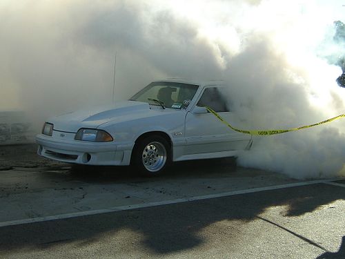 A white Mustang 5.0 automobile performing a burnout on a drag strip