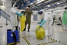 Two people wearing full protective clothing move a plastic trash bag into a marked spot, while their trainer watches them. their trainer.