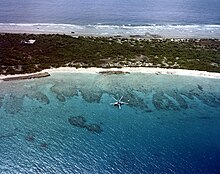 Dark green land with the deep blue sea behind and a lighter blue lagoon in the foreground; the lagoon has isolated reefs visible through its water