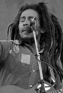 Black and white picture of a man with long dreadlocks playing the guitar on stage.