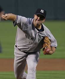 A man in a gray baseball uniform with "New York" across the chest in navy-blue letters and wearing a navy blue baseball cap with an interlocking white "NY" on the front