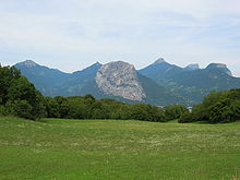 Sloping mountain rising over a meadow, above a valley, with other mountains in the background.