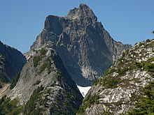 An irregular bare rock mountain peak, with a pronounced shoulder on the left of the picture. In the foreground are two lower prominences, with some trees on them. A snow field is just visible at the foot of the larger mountain.