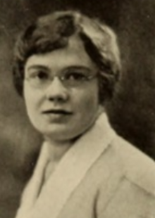 A young white woman with short dark hair, wearing eyeblasses and a light-colored jacket