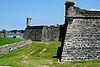 A grassy dry moat separates a low wall on the left from the high walls of the fortress. Made of black stone with white mortar, some of the points of the wall have circular guard towers on them.