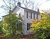 A white house with black shutters and brick chimneys seen from its front left corner. Shrubs and trees obscure the view on the sides.