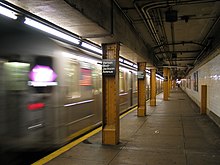 A man wearing a white shirt and blank pants kneels and speaks to a man in a blue shirt who is pinned between a subway train car and a platform. Emergency equipment lies in front of them, while the obscured figures of firefighters and emergency personnel stand behind them.