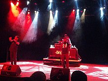 Two African-American men rap into microphones on stage, as a disc jockey plays music in the background