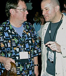 Jonathan Ive is pictured to to the right, talking to John Lasseter who is pictured to the left. The Apple logo can be seen in the background.