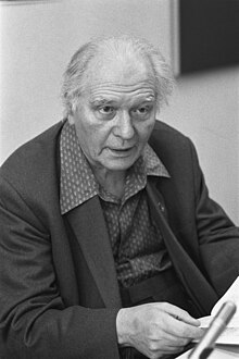 An elderly, balding man with swept-back hair, wearing a suit. He faces the camera.