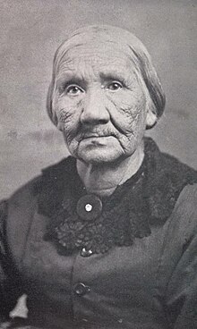 A portrait of an elderly woman with short hair worn back wearing a buttoned dress with a collaret.