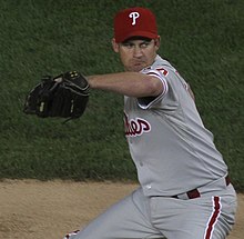 A man wearing a gray baseball uniform and red baseball cap throwing a baseball with his right hand