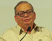 Photograph of a man wearing glasses and speaking in front of a mic.