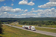 A divided expressway curves from the right of the image through a mostly wooded landscape towards a ridgeline at the rear below a blue sky filled with little white fluffy clouds. Along the roadway closest to the camera is a large white truck with "Perry's Ice Cream" along the side and pictures of scoops of ice cream in various colors and flavors