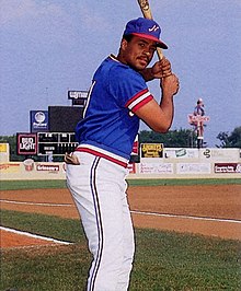 A man wearing a blue baseball jersey with red and white trim, a blue cap with a white "N" on the center, and white pants stands holding a baseball bat preparing to swing.