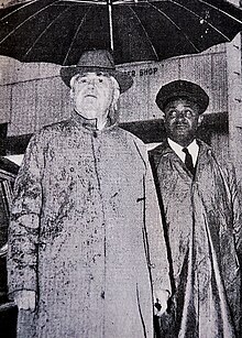 John Lewis in topcoat and hat under umbrella held by James Lewis Jr. in chaufeur uniform.