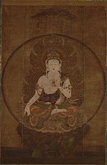 Frontal view of a deity seated on a pedestal in lotus position embellished with ornaments. The right arm is hanging down supported by the legs with the palm of the right hand facing forward. The left hand is placed in front of the body.