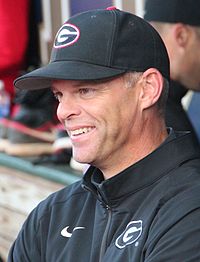 A man wearing a black jacket and baseball cap, both bearing a red and white "G", smiles in a baseball dugout.