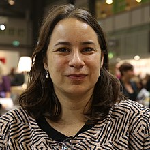 Photography of Ana Martins Marques, a white woman with shoulder-length brown hair, wearing a squared spiral patterned blouse, in a book fair.