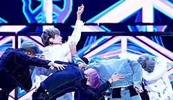 Seven members of BTS performing on stage wearing blue denim jackets. Jin, wearing a white denim jacket, has his hand lifted up in air.