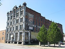 A large, red-brick building with a peeling, white-painted front: A sign on the side of the building is obscured by trees, but partially reads "The Bissman Comp..."
