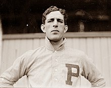A black-and-white photograph of a dark-haired man in a white baseball jersey standing with arms akimbo