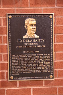 A bronze-and-black metal plaque hung on a brick wall displays an engraving of a man's face; the main caption of the engraving reads "Ed Delahanty; outfielder; Phillies 1888–1889, 1891–1901