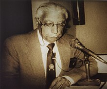 Gulshan Lal Tandon during a meeting wearing a blazer in front of a microphone.