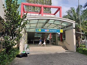 Photograph of station entrance at ground level, encased in a red cubic structure and located in front of a shopping complex.