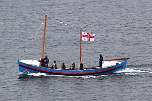 A small blue boat seen from the side. It has a mast at the front and a mast in the centre from which a white and red RNLI flag is flying.