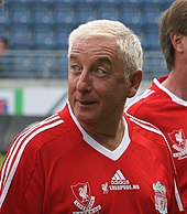 The head and upper torso of a gentleman in his late 50s. He has short white hair and is wearing a red football shirt, which has the 利物浦足球俱樂部 crest on the left breast, and a crest on the right breast that says "利物浦 Legends". A white logo of the Adidas sponsor is visible in the centre of the shirt, and three white stripes are present on the shoulder.