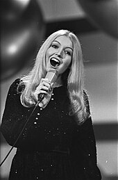 A young woman with long blonde hair wearing a dark dress, singing into a microphone