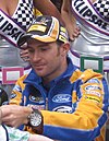 A man aged 30 at an autograph session. He is wearing a black, white and orange baseball cap, a blue and orange fleece and a watch on his left wrist