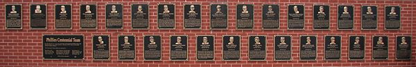 Forty black metal plaques are mounted on a brick wall. They are inscribed with gold images of human faces and text.