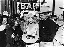 梦露于《乱点鸳鸯谱》。她拿着装满美元钞票的宽沿帽，旁边站着克拉克·盖博和塞尔玛·里特尔（Thelma Ritter）。他们身后是一个写着“BAR”的标志和一群人。