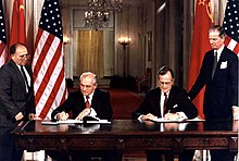 Two men in suits sit signing documents at a large table in front of their countries' flags while two others stand outside watching them.