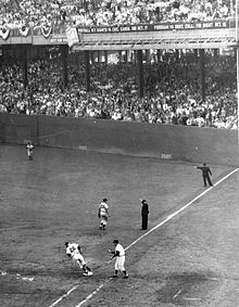 A black and white image of right field and first base in a baseball diamond in a large stadium with filled stands. A runner is rounding first