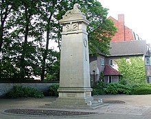 A four-sided stone column topped with a lion lying down, in a small semi-circular paved garden area.