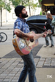 Jay Sparrow standing on the street, wearing dark blue jeans, a blue-and-white checkered shirt, and a black knit tuque, playing an acoustic guitar and singing