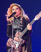 嘎嘎小姐手拿粉红吉他，站在麦克风的后面，穿着黑色皮革流苏衣。