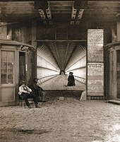 A square opening forms the entrance to the bridge's lower level. Vertical wooden supports line the level. Flagstones pave the entrance and a booth stands to the left. A man stands at the booth, another sits in front of the entrance, and another is walking up the stairs on the right.